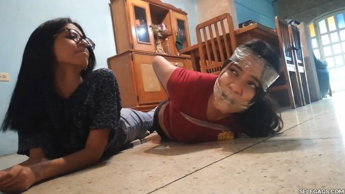 Gagged girl hogtied by girl with glasses in tight tape bondage