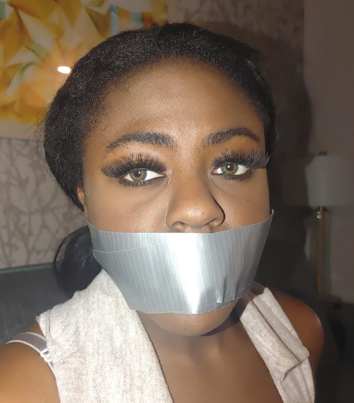 A black woman with green eyes is gagged with silver duct tape in a living room