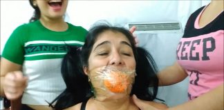 South American MILF Turned Gag Slut A woman with pigtail hair has her mouth gagged with a rubber ball and transparent tape wrapped around her head. Two women are standing behind her.