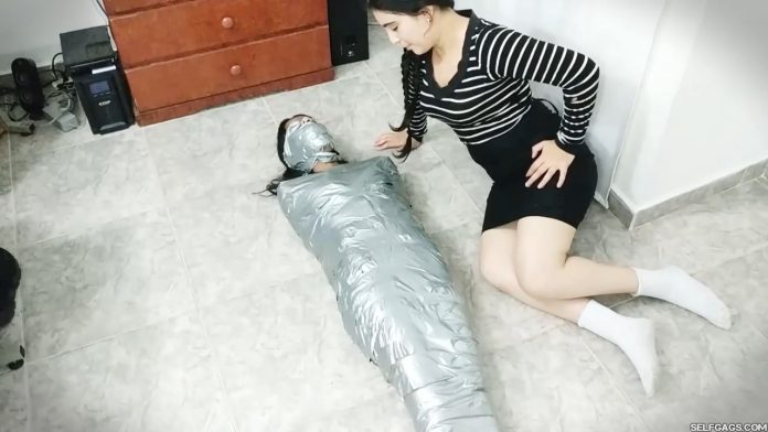 A girl is mummified, gagged and blindfolded with duct tape by a girl dressed in a costume as Wednesday Addams