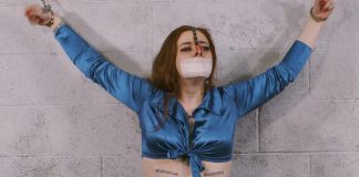 Freshie Juice Gagged & Nose-Hooked for Game Day Control Handcuffed girl with red hair is wearing a medical tape gag and a nose hook pulling her nose upwards