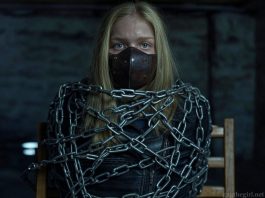 Blonde teen Everly, muzzled with a thick brown leather BDSM gag, heavily shackled in steel chains, wearing a black leather jacket, sitting helplessly in a dark basement.