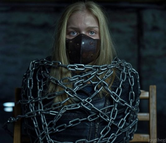 Bound Beauty Everly: Shackled, Gagged, and Helpless in Leather Blonde teen Everly, muzzled with a thick brown leather BDSM gag, heavily shackled in steel chains, wearing a black leather jacket, sitting helplessly in a dark basement.
