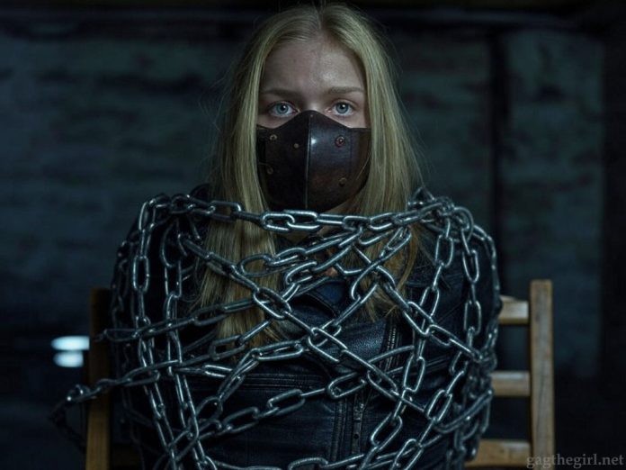 Blonde teen Everly, muzzled with a thick brown leather BDSM gag, heavily shackled in steel chains, wearing a black leather jacket, sitting helplessly in a dark basement.