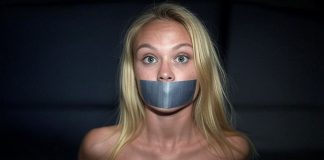 Danish Event Girl Asta Bound and Tape Gagged in Darkness Blonde Danish event girl named Asta, duct tape gagged and bound, wearing a black glittering top, wide-eyed and staring fearfully in a dark setting.