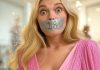 Tape Gagged Shock: Blonde Lila’s 18th in Pink Blonde girl Lila, wide-eyed in shock, wearing a pink dress, duct tape gagged with '18' written on it, in a cozy room.
