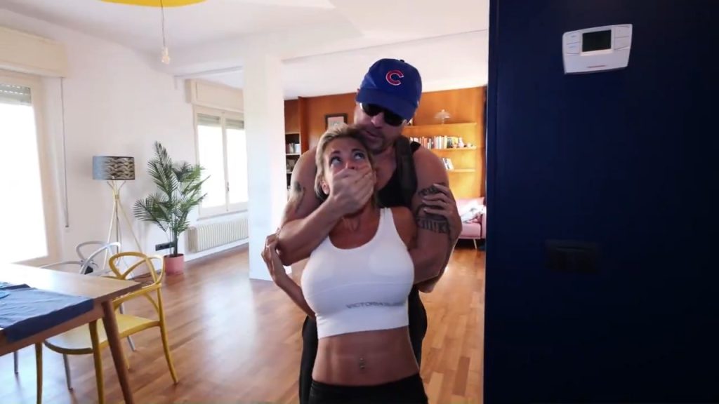 Terrified Elenoire Steiner in white crop top and black dress gets brutally hand-gagged and roughly pinned from behind by fugitive Tony Houston moments after he breaks in, her big breasts pressed tight as he drags her through the apartment – Tony Houston Bondage Paranoia home invasion terror