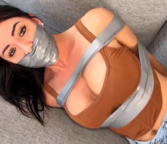 Carlita Cruz First Time: Mouth Stuffed & Tape Sealed by Neighbor – Silenced Cuties Carlita Cruz lying on couch in tight duct tape bondage, gray tape gag sealing her lips, big tits wrapped and exposed - Silenced Cuties