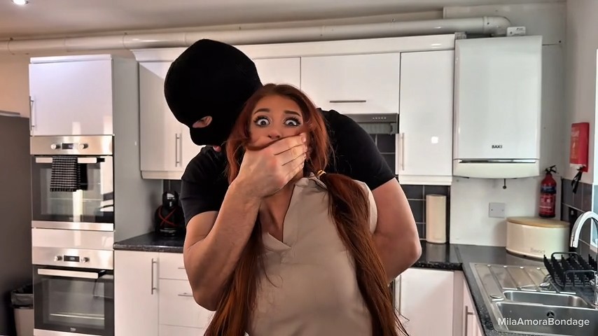 Masked intruder grabs red-haired woman from behind in kitchen, hand over her mouth, eyes wide in surprise - MilaAmoraBondage studio.