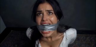 Woman with duct tape over her mouth, looking upward with a distressed expression in a dim room.