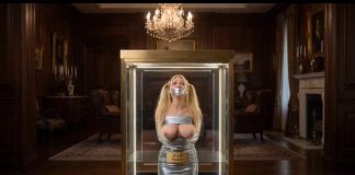 Wide shot of the glass display case elevating with bound and gagged teacher Julie Millard inside opulent room