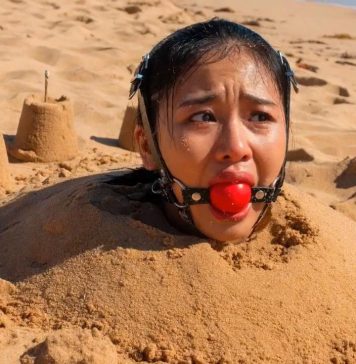 Southeast Asian Harness Ballgagged & Bucket Hooded on Beach – AI Bondage Close up of terrified Southeast Asian beauty with thick harness ball gag strapped in her mouth, eyes wide with fear