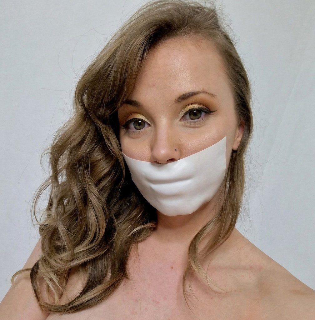 Lil Missy UK with a tight white microfoam tape gag pressing against her lips, looking at the camera in a close-up portrait