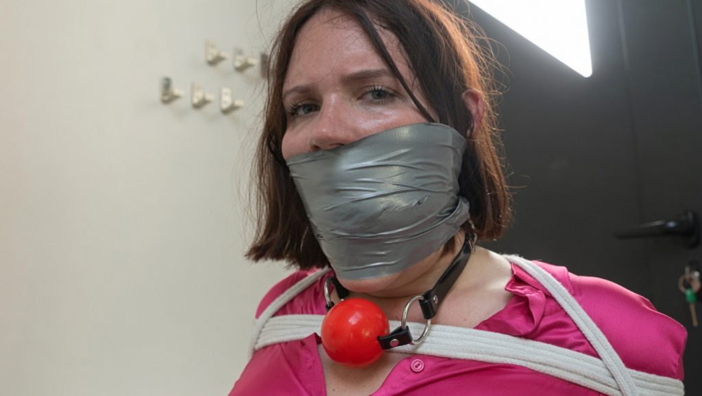 Snowflake from FrenchBondage with a thick silver duct tape gag tightly sealed over her mouth, looking strained, with a red ball gag hanging around her neck