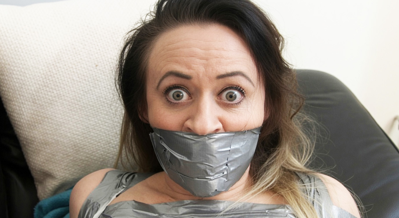 Brunette woman with wide eyes tightly gagged with multiple layers of silver duct tape covering her mouth and lower face