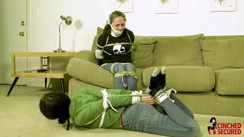 Living-room dual — skull-shirt captive in a thick white cloth cleave on the olive-green sofa with thigh/ankle rope; green-shirt partner hogtied on beige carpet