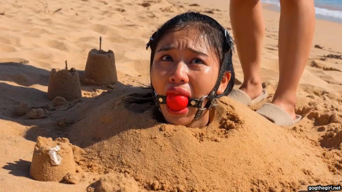 Unknown woman in slippers approaching the harness ballgagged Southeast Asian girl buried in the sand