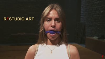 Closer look at Lali looking toward the camera, gagged, harness pulling her elbows back