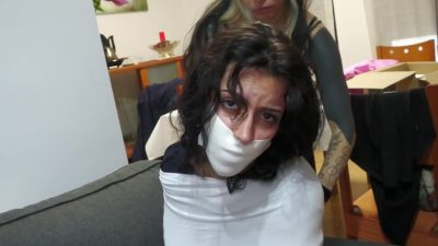 Medium: Persefone already microfoam-tape-gagged looks to camera while Liricaa wraps more white microfoam around her torso