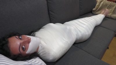 High wide: finished full-body microfoam mummy on the grey sectional, bare feet out, eyes to camera with white tape gag sealed