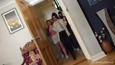 Ungagged ID — Eliza (pink tee, white shorts) and Roxxi (cream blazer, purple updo, necktie) stand hands-up in the doorway while the skull-masked Serpent escorts them in