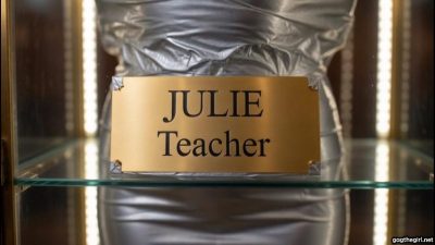 Close-up of brass plaque reading JULIE Teacher on silver duct tape mummified body in glass display case