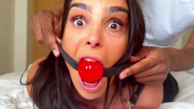 Big Back D's hands buckling a bright red ball gag into Latina's mouth on the white bed