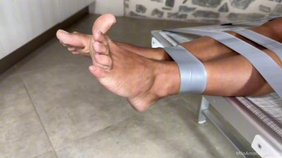 Close of Alba’s bare feet with silver duct tape wrapping both ankles to the lounger frame