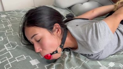 Tight face close-up of Barby’s bright red ball gag and black leather strap on the grey tee