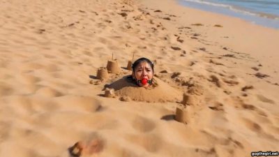 Southeast Asian woman buried neck-deep in sand wearing tight black harness ball gag on deserted island beach