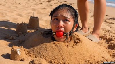 Unknown woman in slippers approaching the harness ballgagged Southeast Asian girl buried in the sand