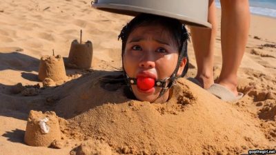 Unknown female slowly lowering bucket over the head of the harness ballgagged woman buried on the beach
