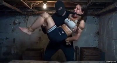 Masked man carrying struggling ballgagged sporty girl in fireman’s carry inside dirty basement with exposed pipes