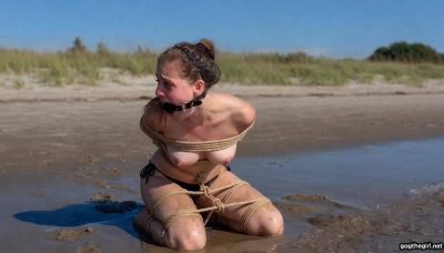 Side view of tearful ballgagged university student struggling desperately in tight rope bondage on wet sand at the shoreline