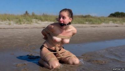 Distressed ballgagged student kneeling in tide with heavy chest and crotch ropes, ocean waves around her bound body